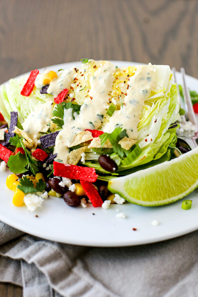 A wedge of iceberg lettuce with corn, black beans, cilantro, green onion, a lime wedge, queso fresco, tortilla strips and creamy chipotle tomatillo dressing.