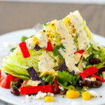 A wedge of iceberg lettuce with corn, black beans, cilantro, green onion, a lime wedge, queso fresco, tortilla strips and creamy chipotle tomatillo dressing.